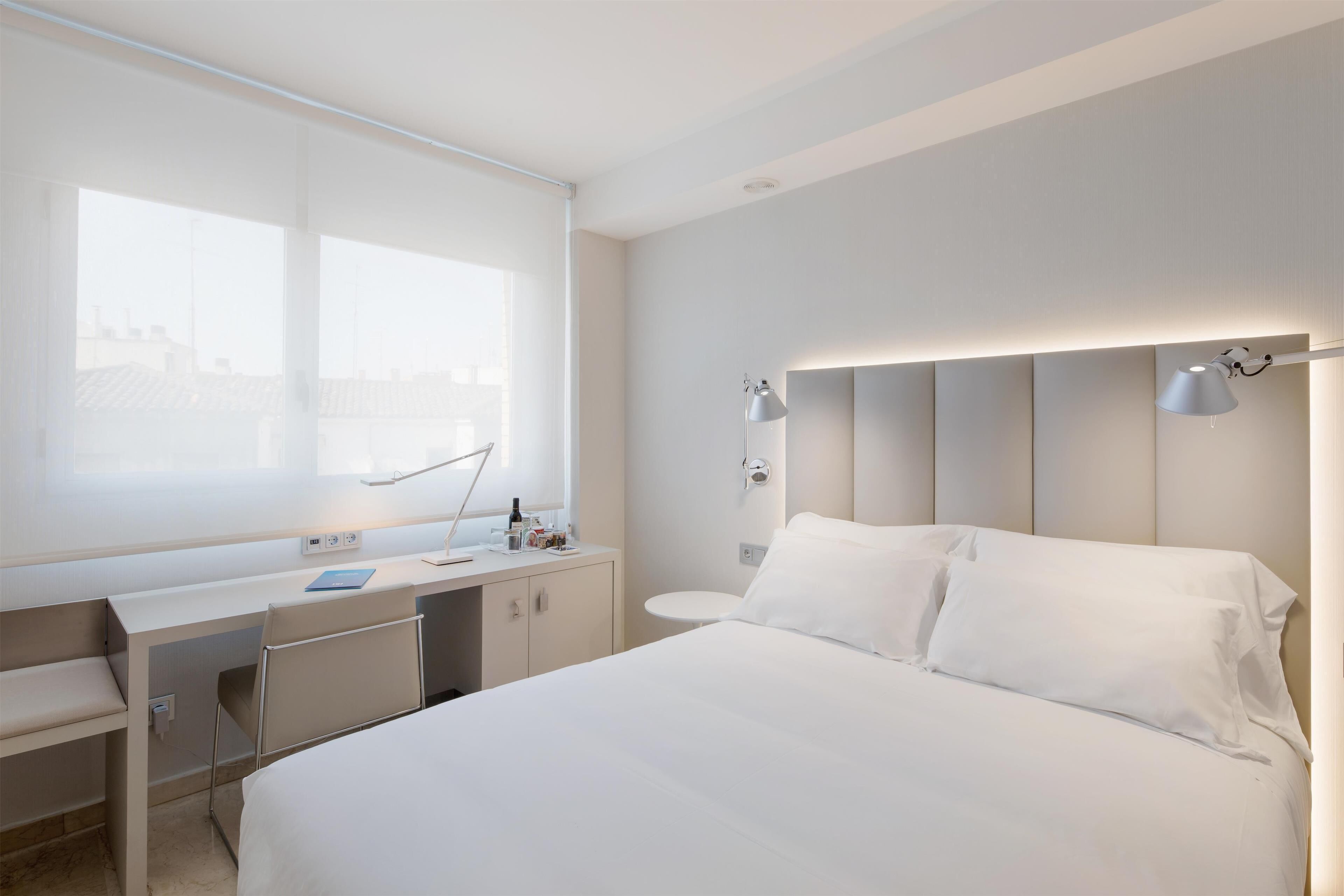 A modern hotel room with a large bed, a desk with a chair, and a window providing a city view.