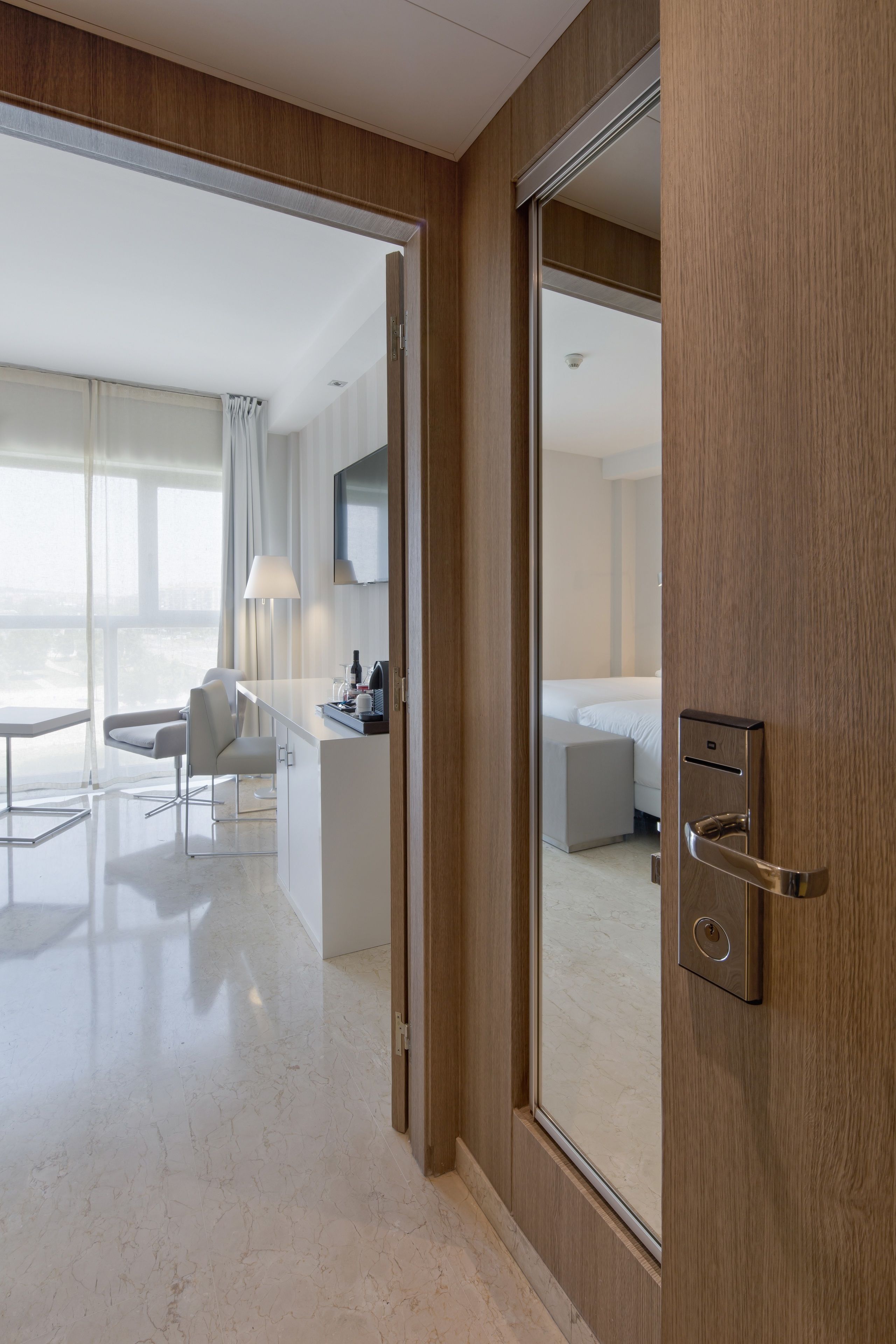 Una habitación de hotel moderna con puerta de madera, cama, escritorio y zona de estar con una silla y una lámpara.