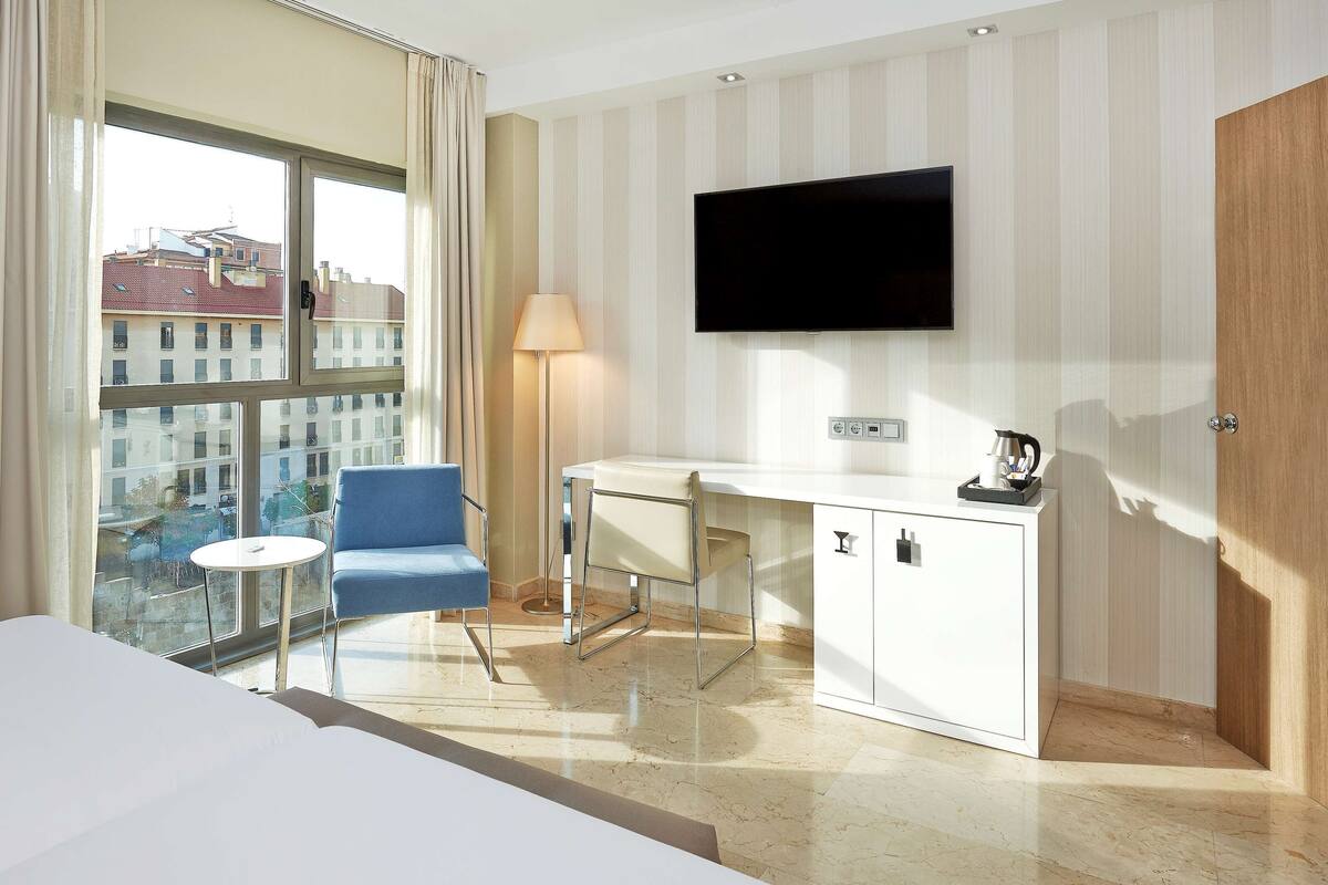 Une chambre d’hôtel équipée d’un lit, d’un bureau, d’une chaise, d’un téléviseur fixé au mur et offrant une vue sur les bâtiments à travers de grandes fenêtres.
