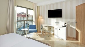 Habitación de hotel con cama, escritorio, silla, televisor montado en la pared y vistas a edificios a través de amplios ventanales.