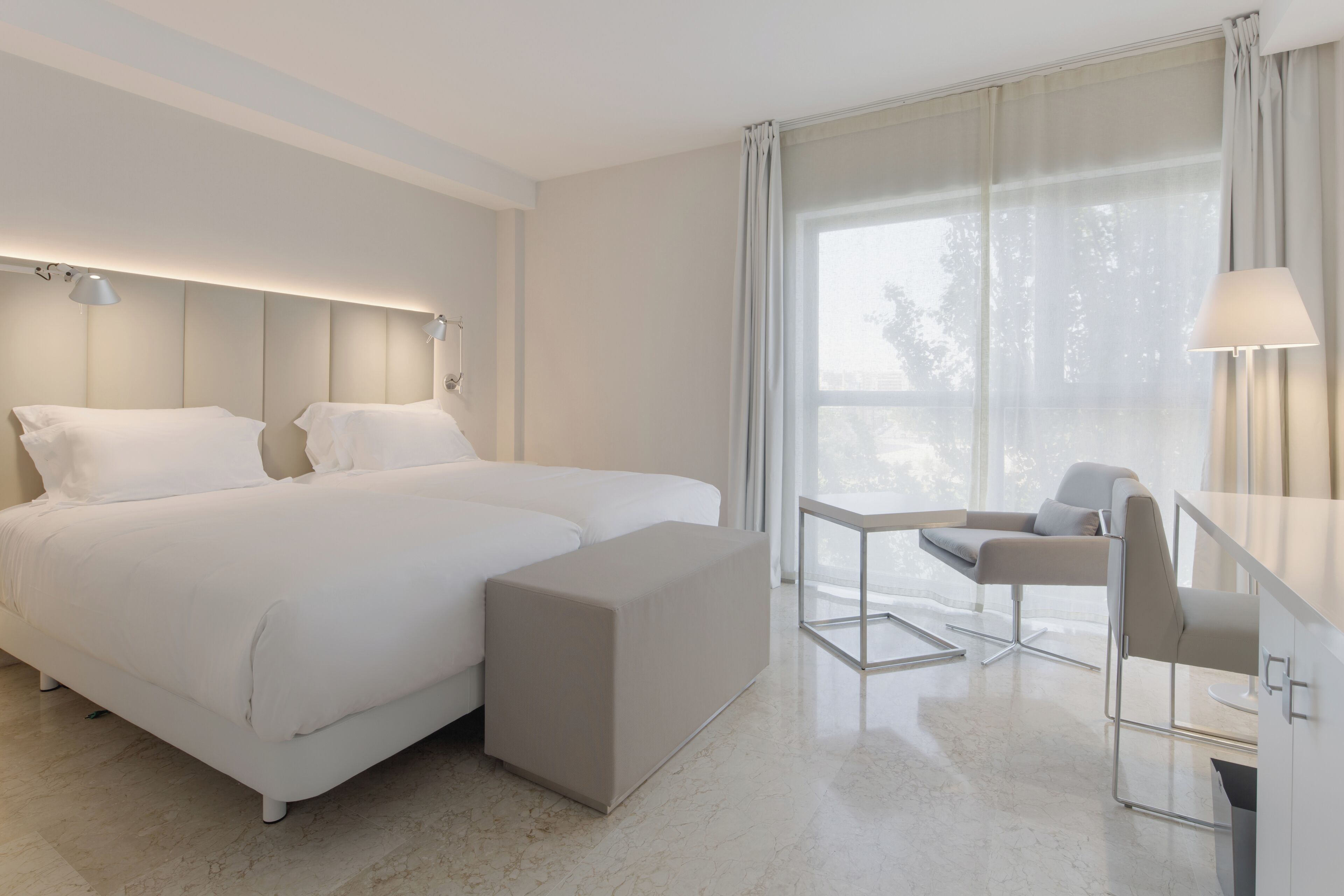 A modern hotel room with a large bed, a small sofa, a desk, and a window with curtains.