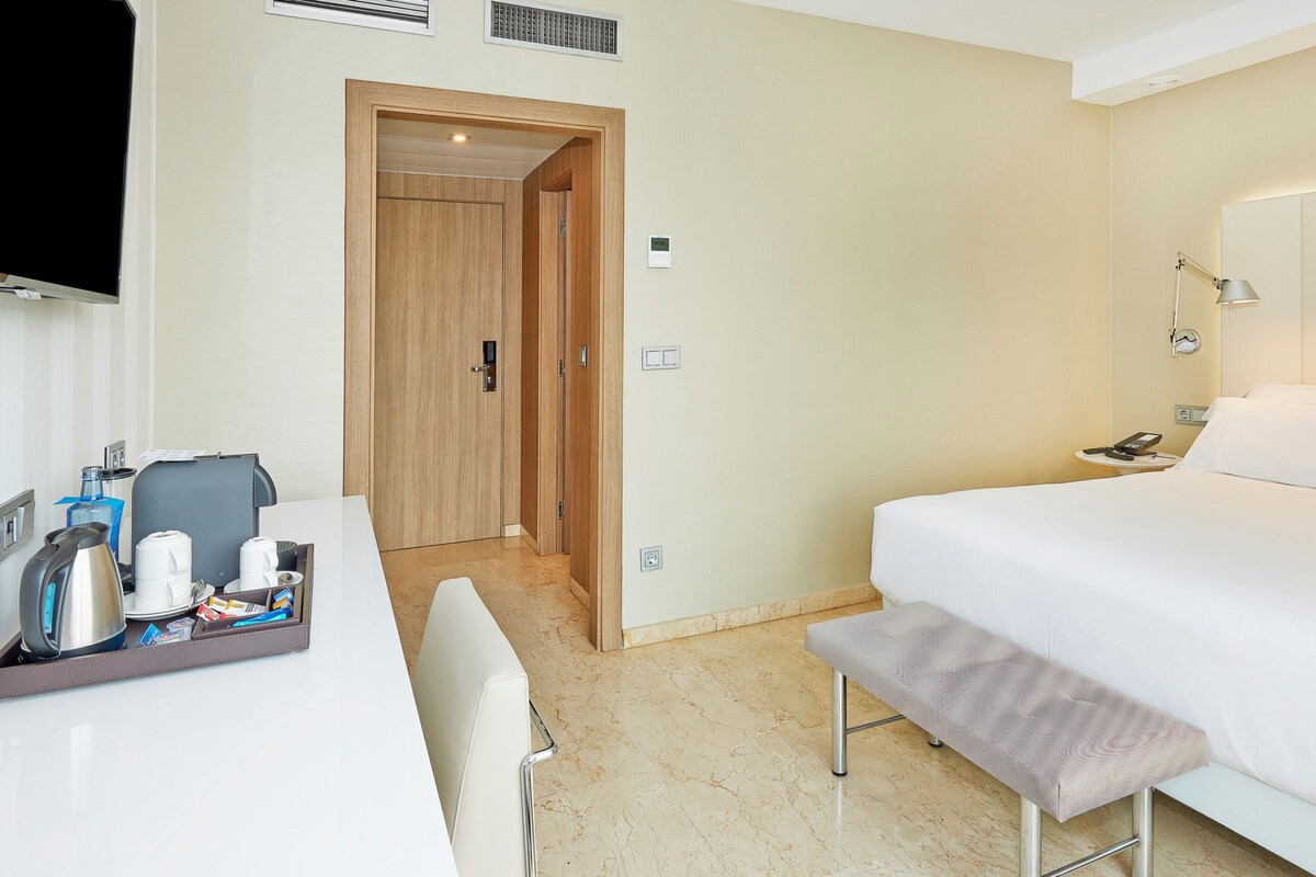 Una habitación de hotel con cama, mesita de noche, escritorio con cafetera y televisor.
