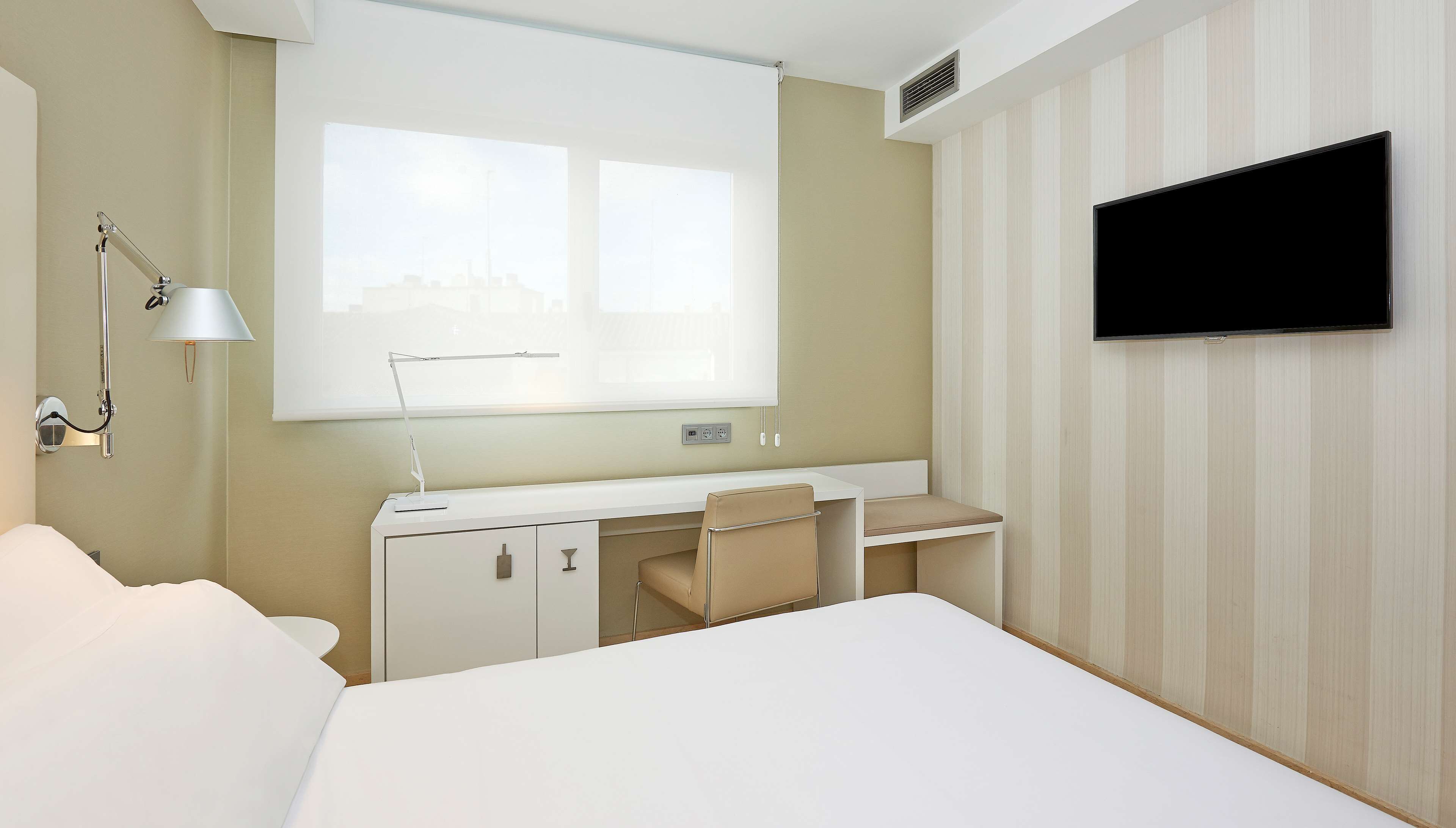 A hotel room with a bed, a desk with a chair, a TV mounted on the wall, and a window with blinds.