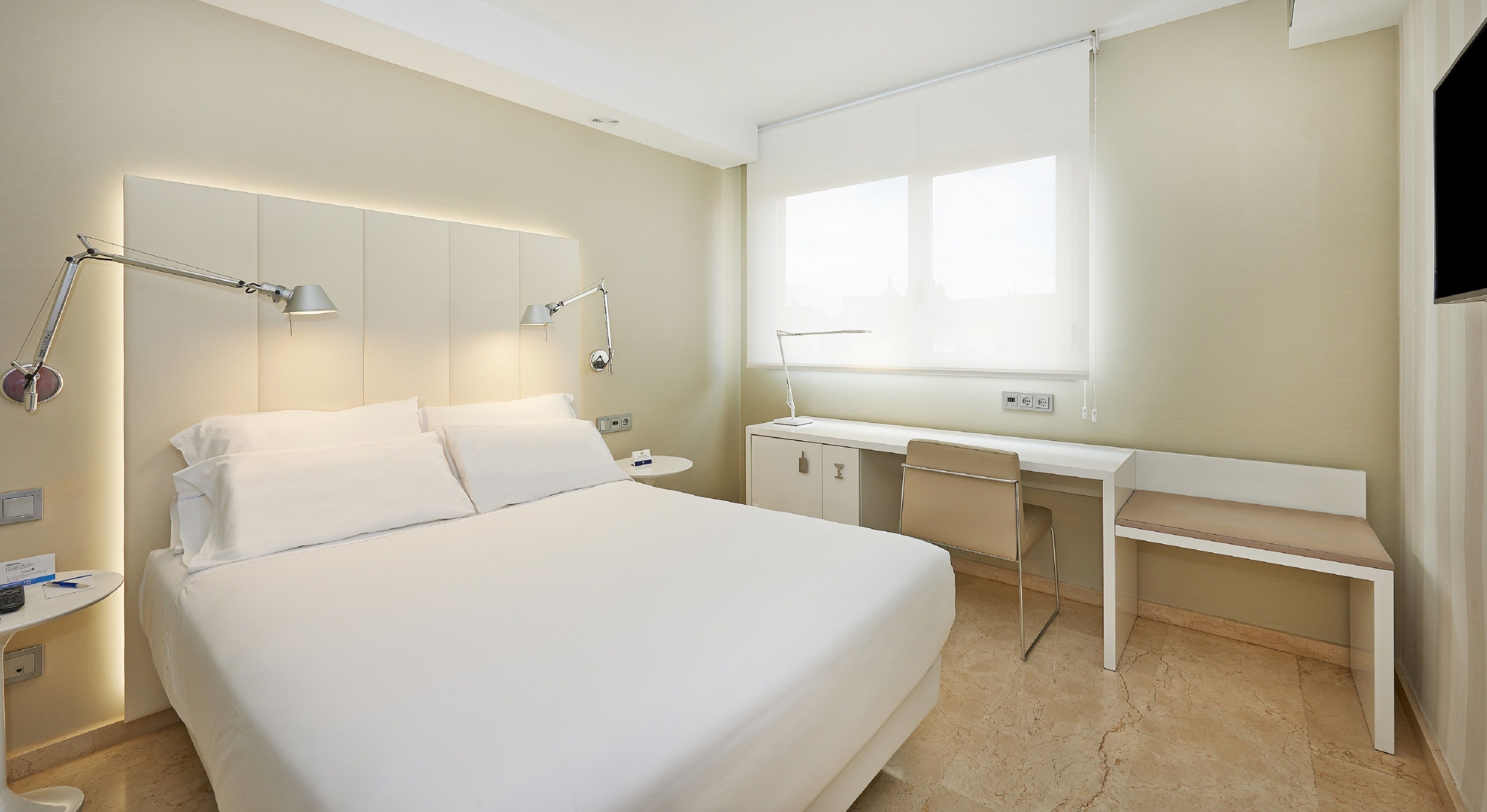 A modern hotel room with a large bed, bedside tables, a desk, and a window.