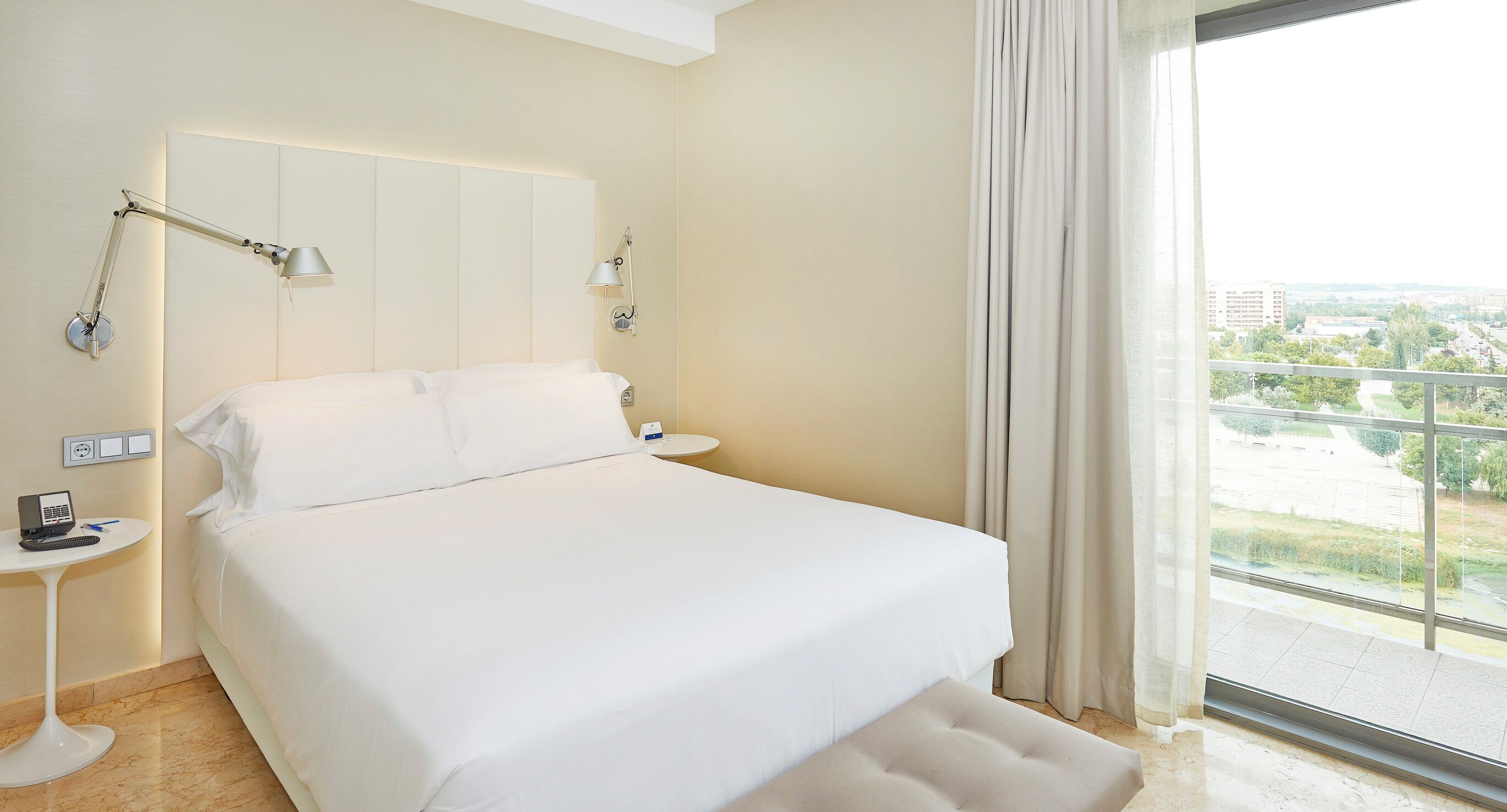 A hotel room with a large bed, two bedside tables, a desk with a computer, and a view of a cityscape through a large window.