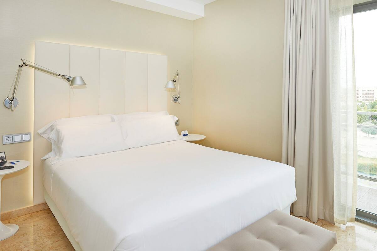 Une chambre d’hôtel avec un grand lit, deux tables de chevet, un bureau avec un ordinateur et une vue sur le paysage urbain par une grande fenêtre.