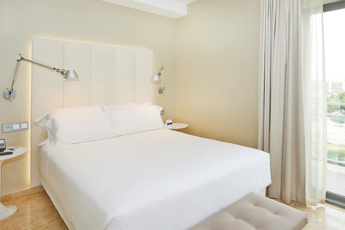 A hotel room with a large bed, two bedside tables, a desk with a computer, and a view of a cityscape through a large window.