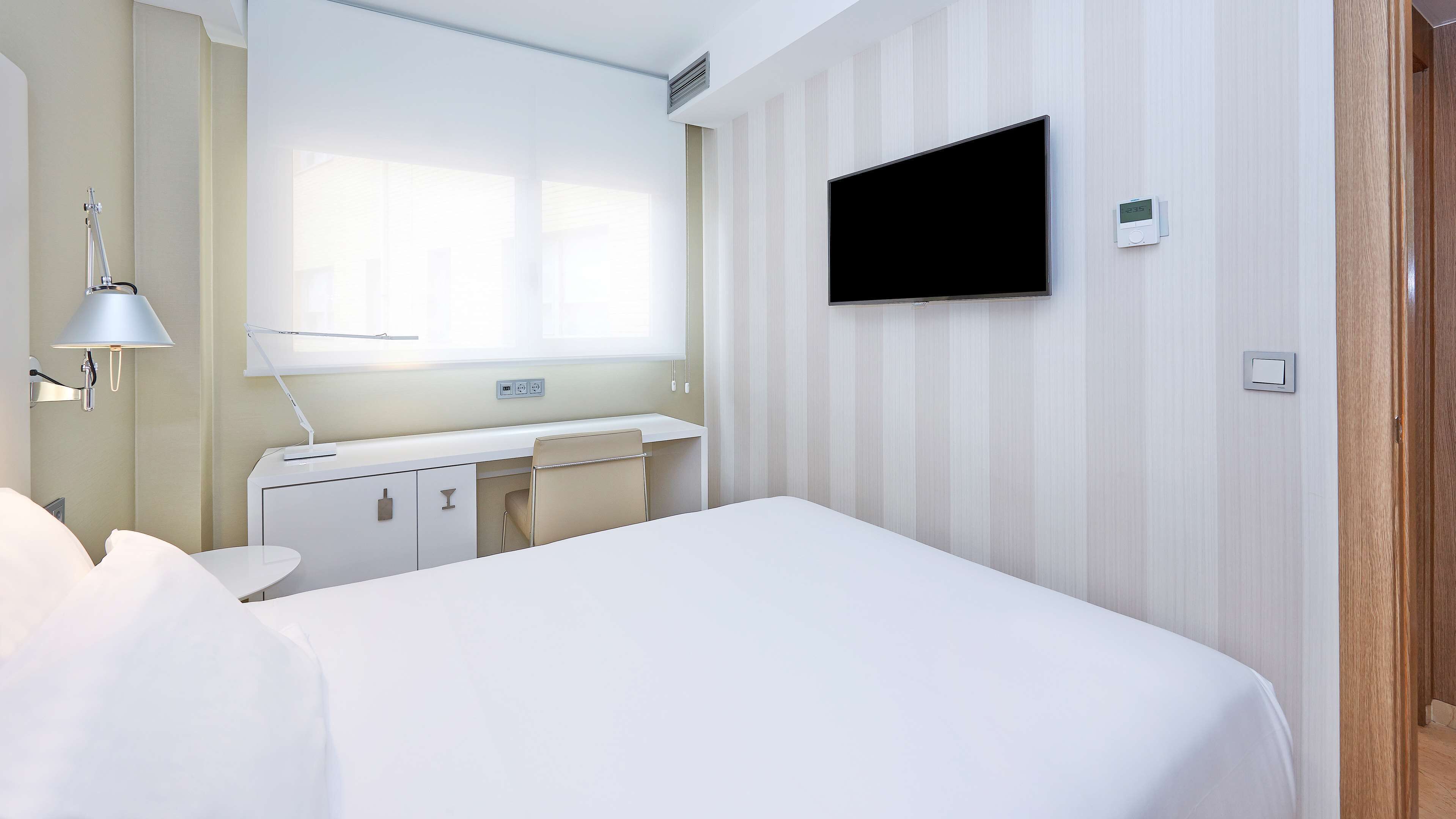 A hotel room with a bed, a desk, a chair, a TV mounted on the wall, and a window.
