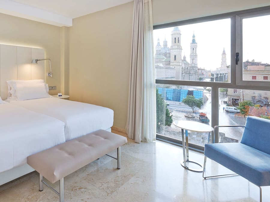 Habitación de hotel con dos camas, un banco, una silla, una mesita y vistas de la ciudad a través de amplios ventanales.