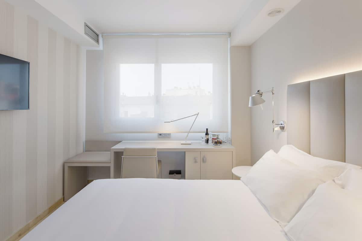 A hotel room with a bed, a desk, a chair, a window, and a wall-mounted lamp.