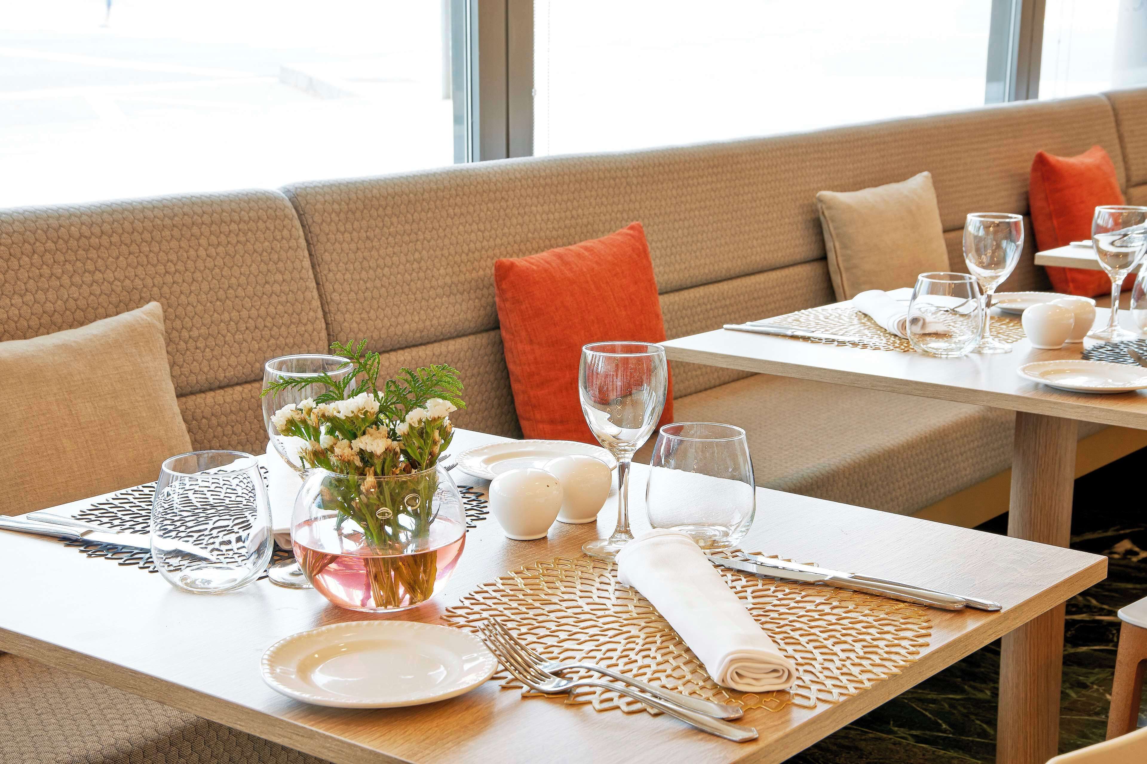 Un comedor con sofá, mesas dispuestas con cubiertos, platos y vasos, y un jarrón con flores.