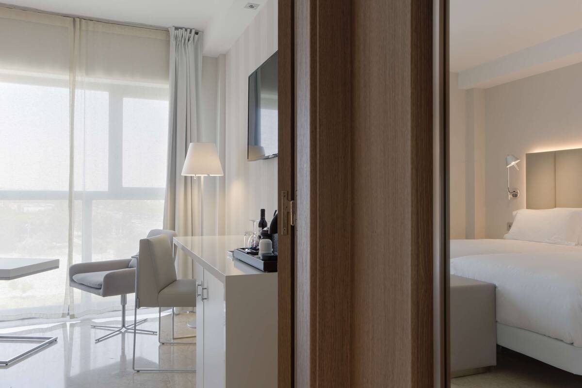 Une chambre d’hôtel moderne, équipée d’un lit, d’un bureau avec une lampe, d’une chaise et d’une grande fenêtre avec des rideaux.