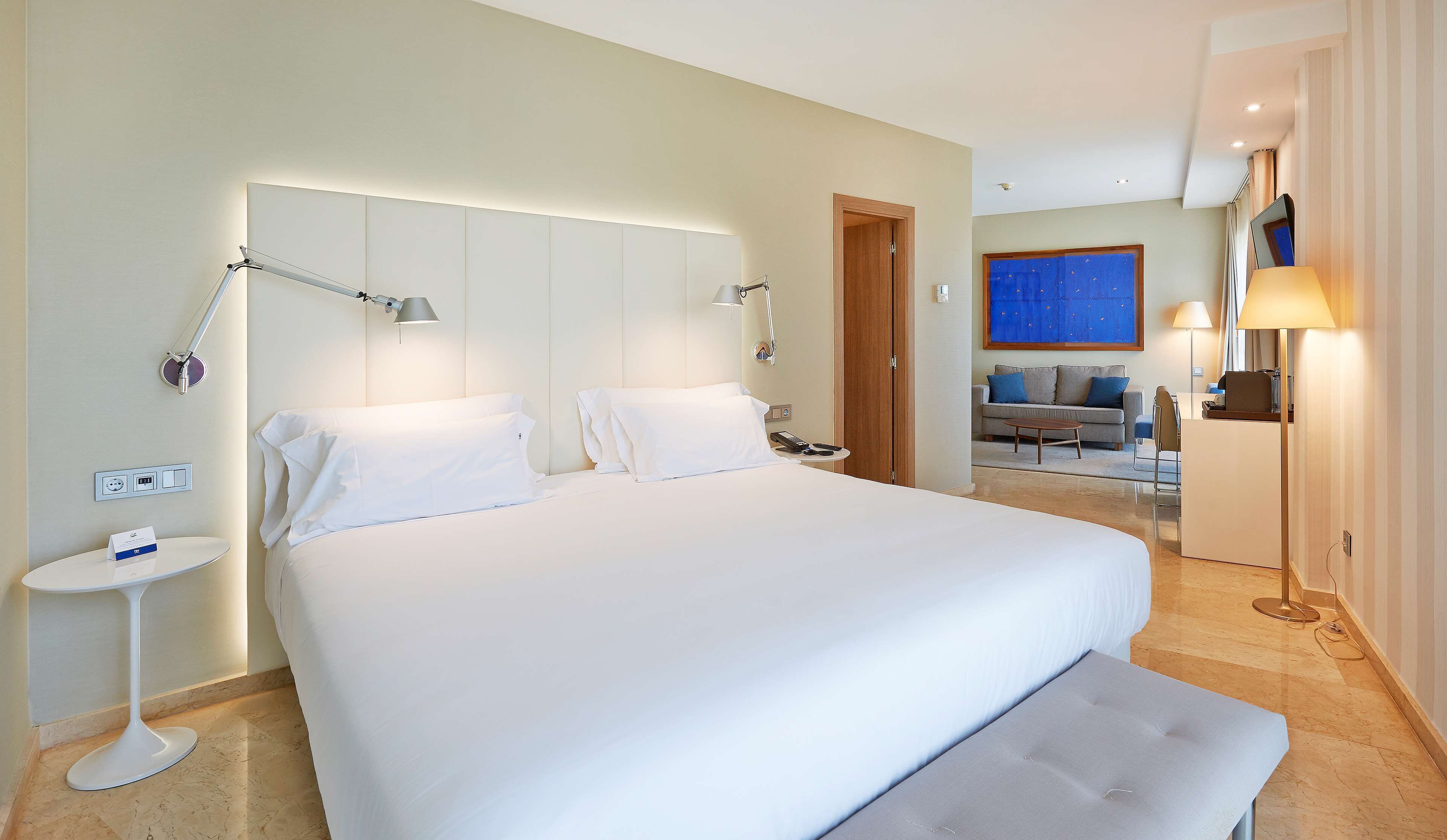 A hotel room with a large bed, bedside lamps, a small table, and a sofa in the background.