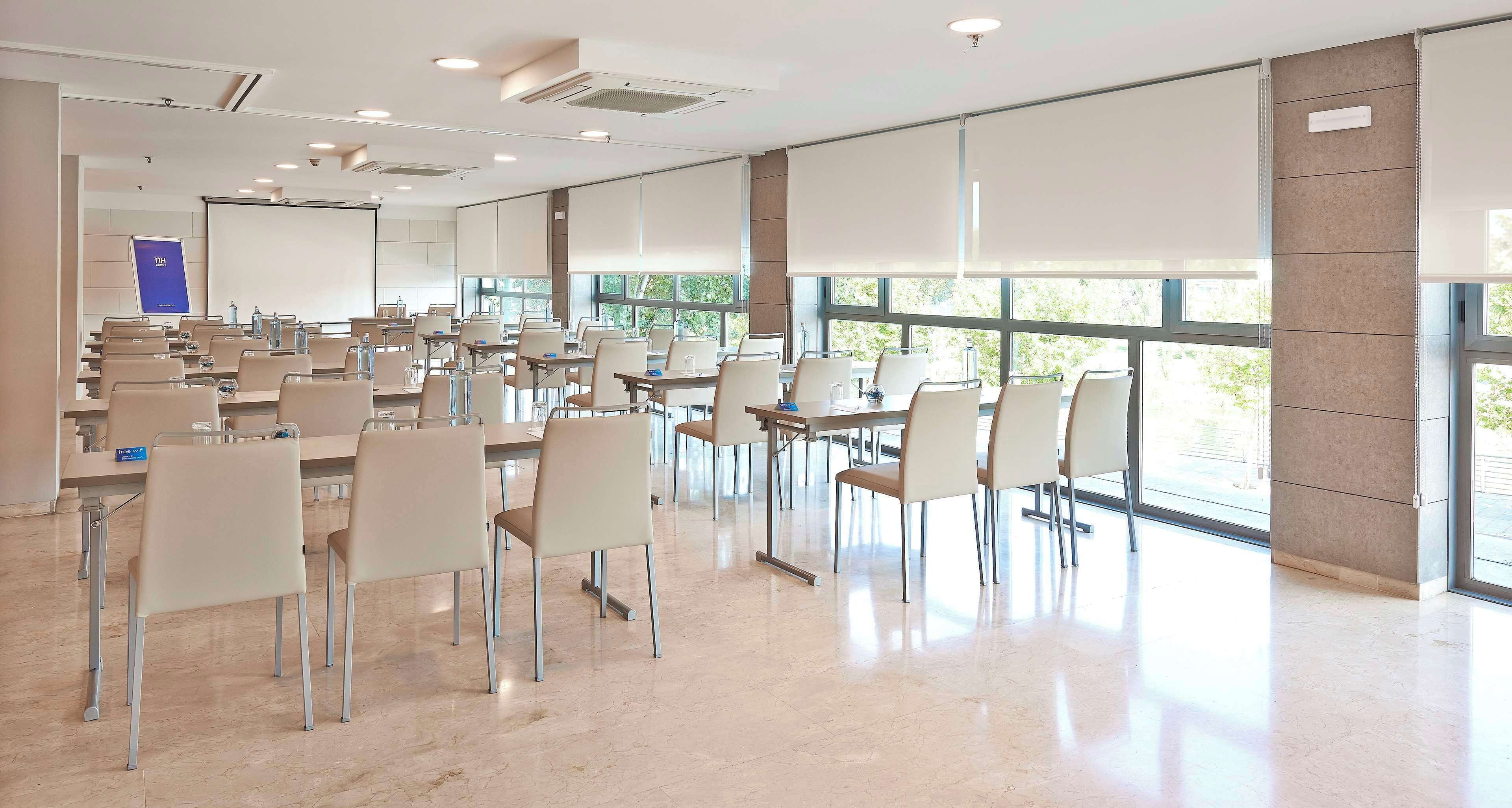Una amplia sala de conferencias con múltiples mesas y sillas, ventanas grandes y una pantalla de proyector.
