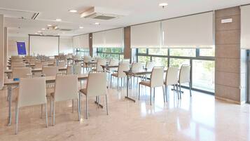 Una amplia sala de conferencias con múltiples mesas y sillas, ventanas grandes y una pantalla de proyector.