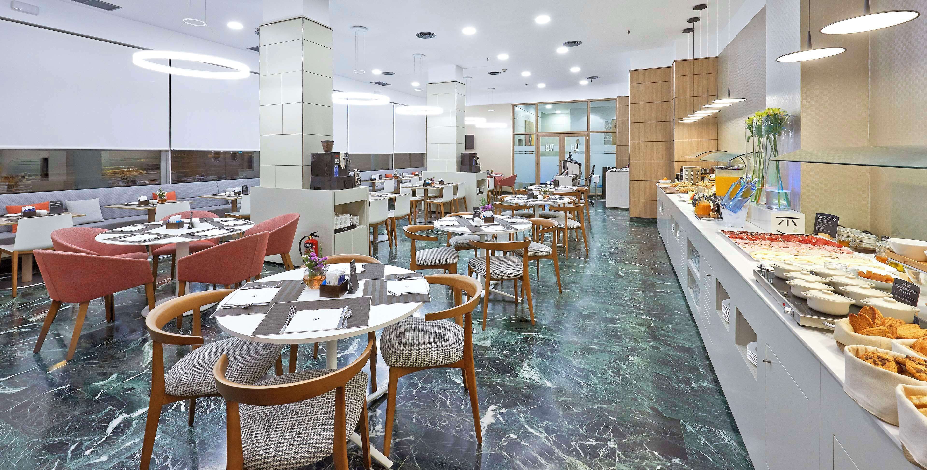 Un restaurante moderno con mesas y sillas redondas, un mostrador de buffet con diferentes opciones de comida y un amplio ventanal con vistas a la calle.