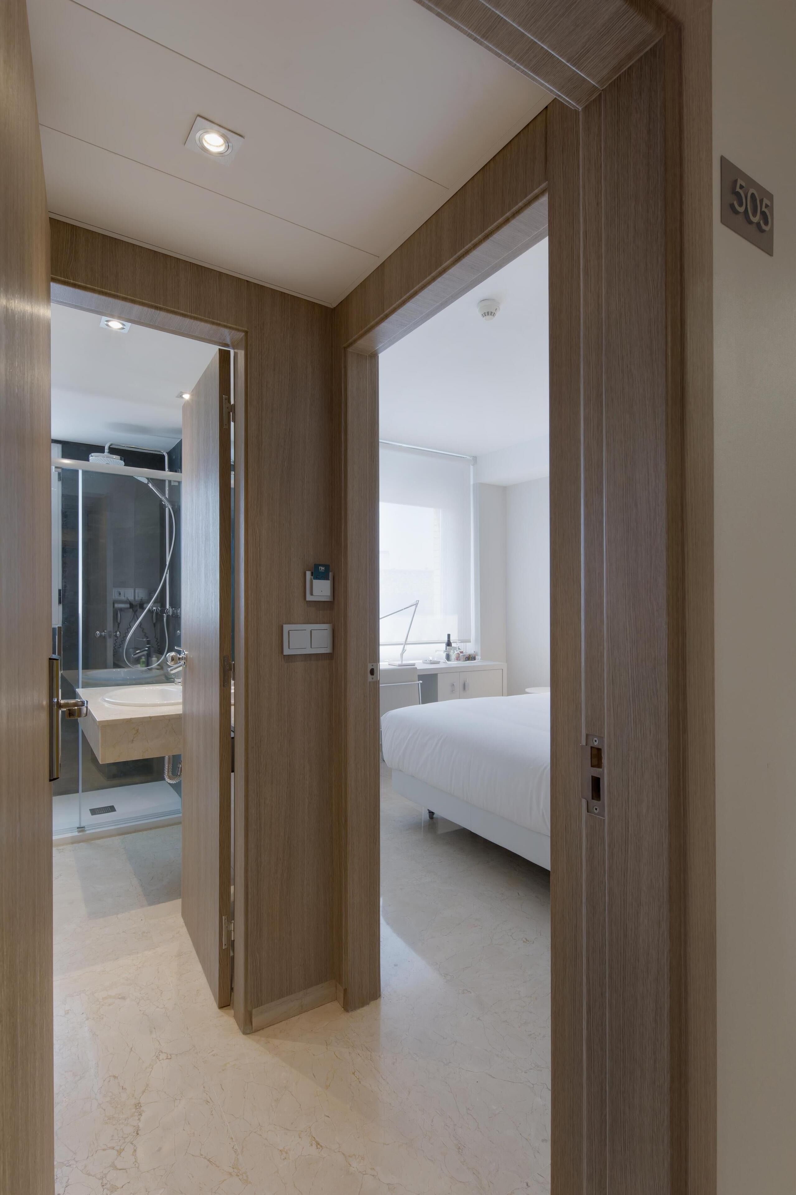 Una habitación de hotel con puerta de madera, cama, mesita de noche y ventana.