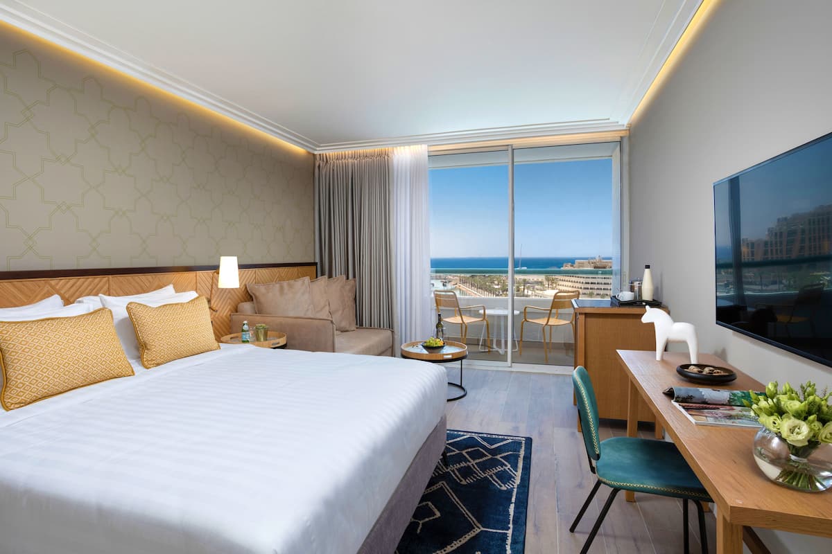 Ein Hotelzimmer mit einem großen Bett, einem Schreibtisch, einem Sessel, einem Fernseher und einem Balkon mit Meerblick.