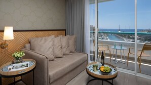 A modern hotel room with a sofa, a small round table with a fruit bowl, a lamp, and a view of the ocean and marina.