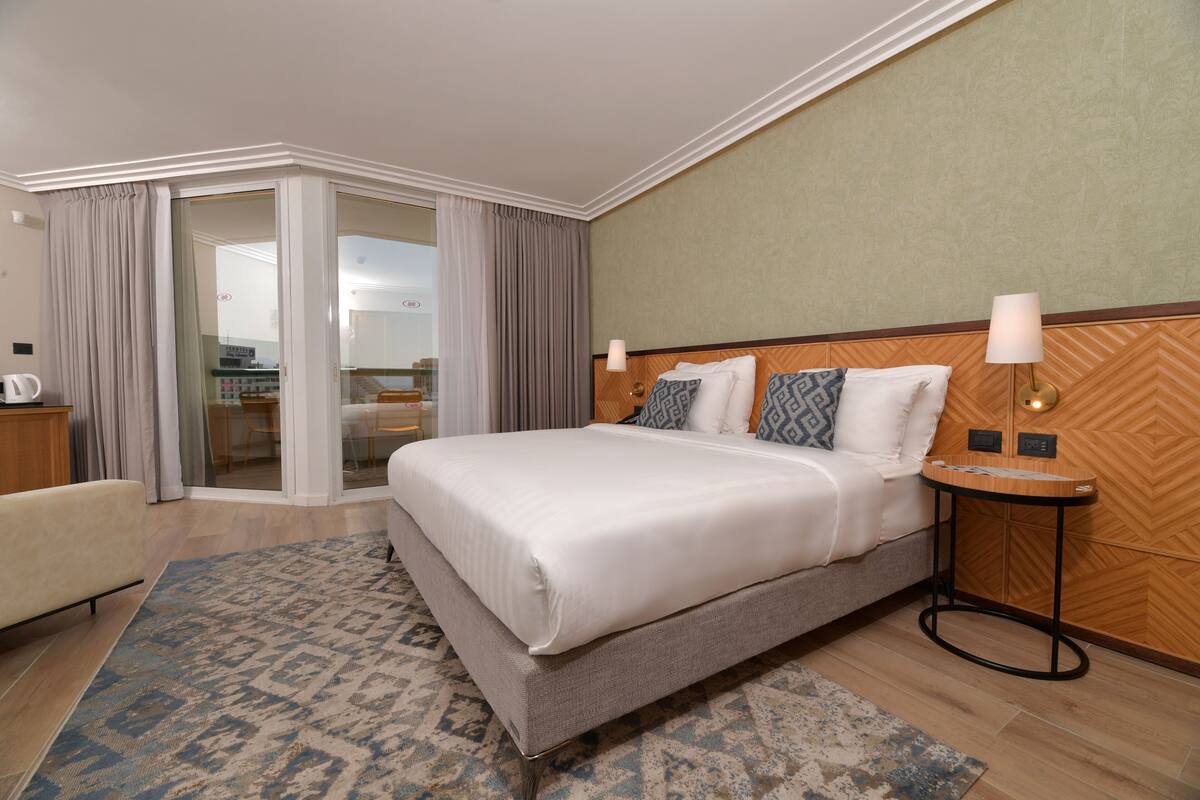 Ein Hotelzimmer mit einem großen Bett, einem Nachttisch, einem Sessel und Blick ins Freie durch eine Schiebetür.