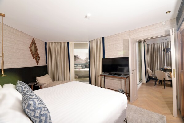A modern hotel room with a large bed, a desk with a flat-screen TV, a dining area with chairs, and a view of the city through the window.