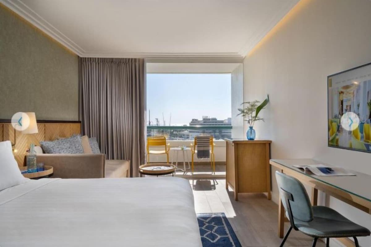 Quarto de hotel com cama, escrivaninha, cadeira, mesa pequena, luminária, vaso com planta e vista da cidade por uma janela grande.