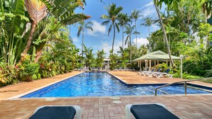 Una piscina con sillas de descanso y un área cubierta, rodeada de plantas tropicales y palmeras.