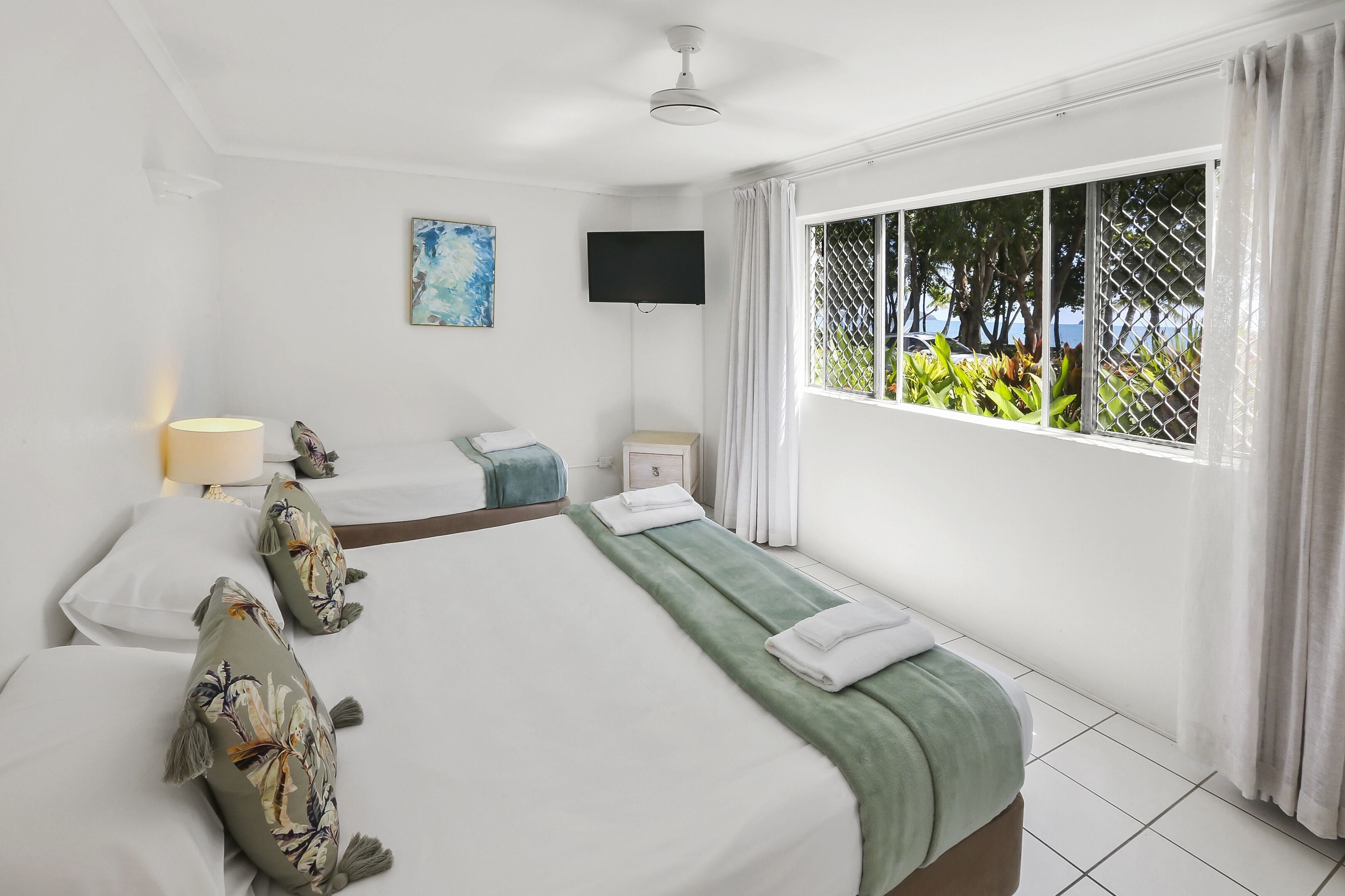 A hotel room with two beds, a flat-screen TV, a window with a view of greenery, and a ceiling fan.