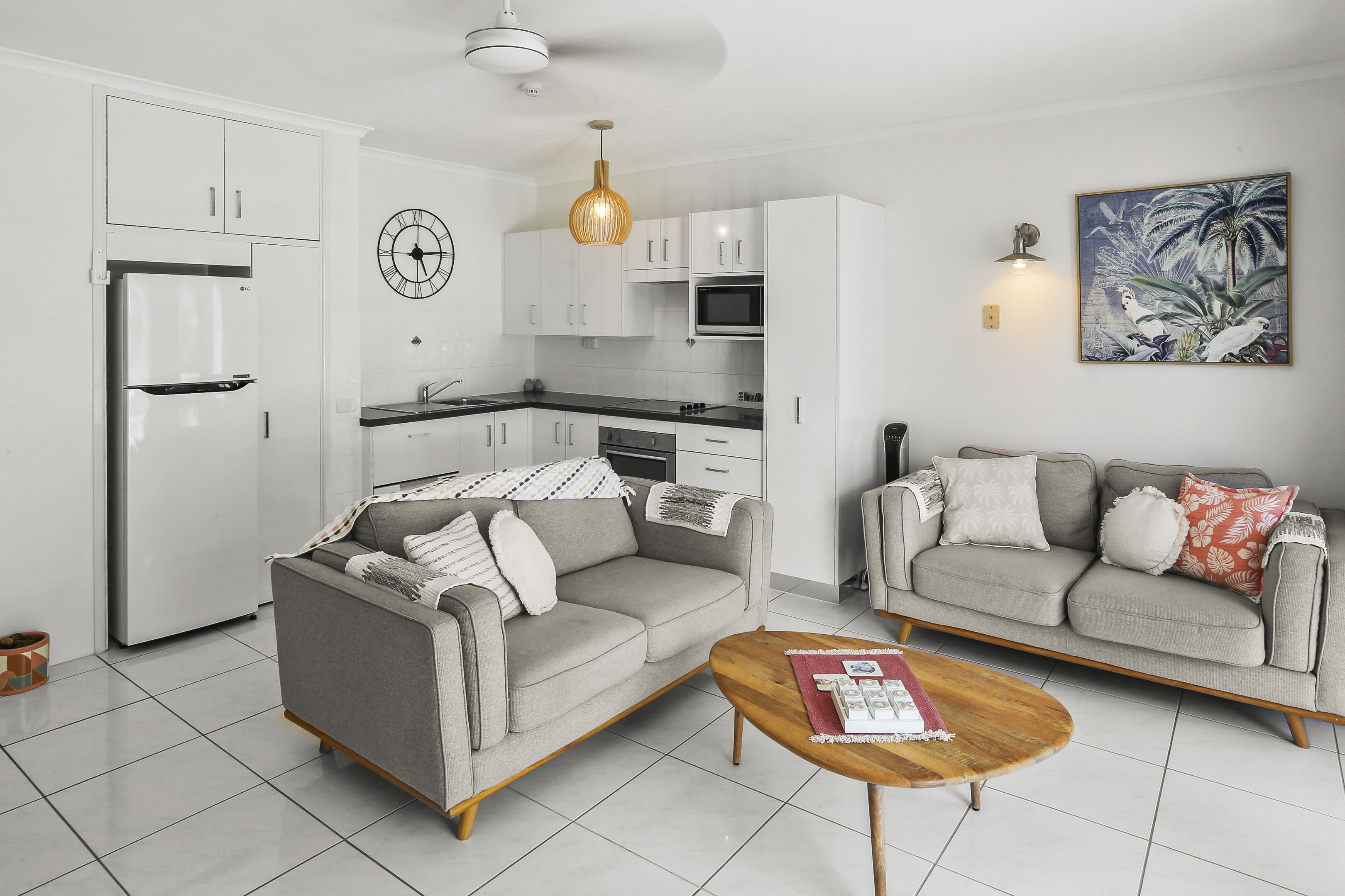A modern kitchen with white cabinets, a refrigerator, and a microwave. There is a gray sofa with a wooden coffee table in the living area.