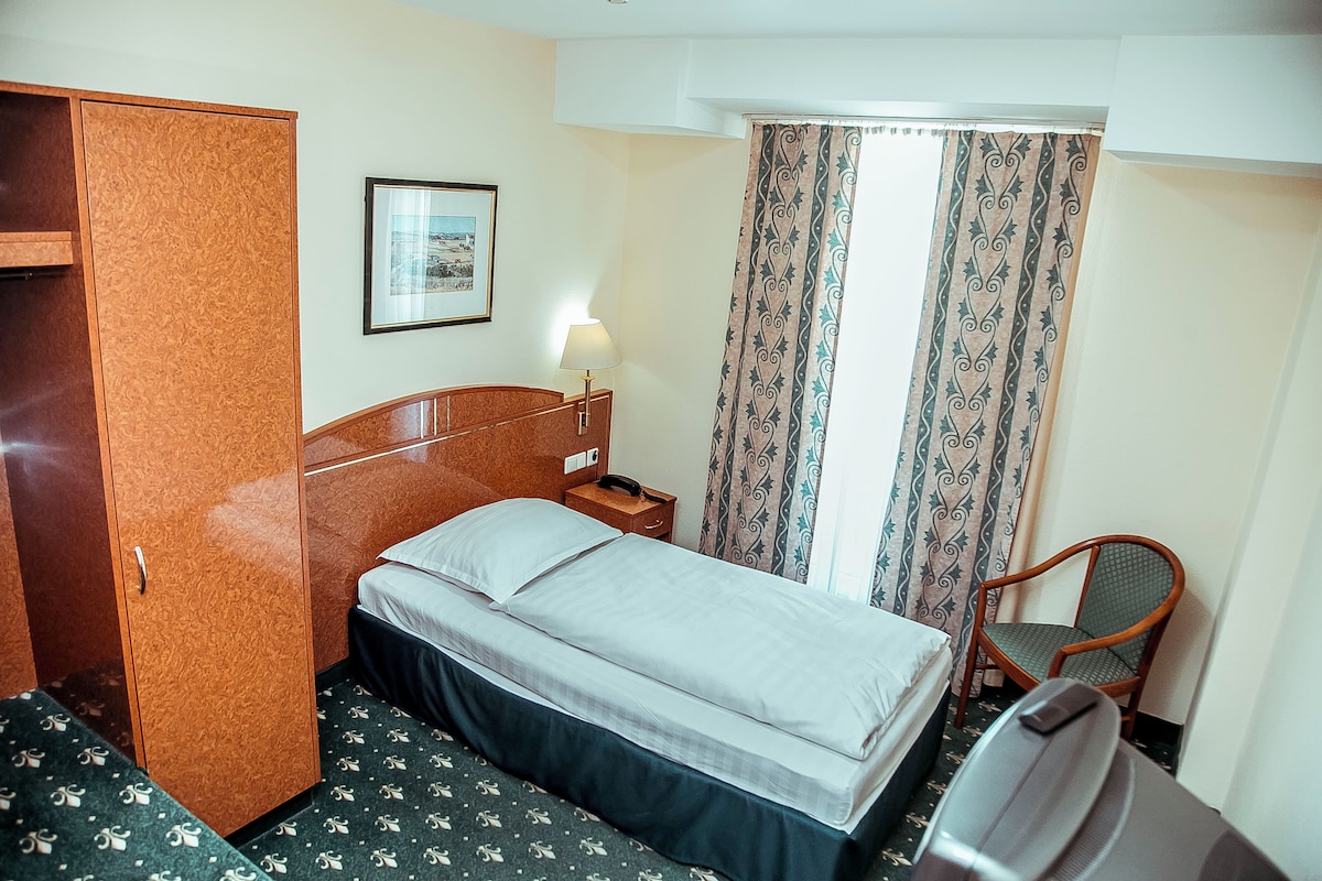 A hotel room with a bed, a wooden wardrobe, a chair, a bedside table with a lamp, and a framed picture on the wall.