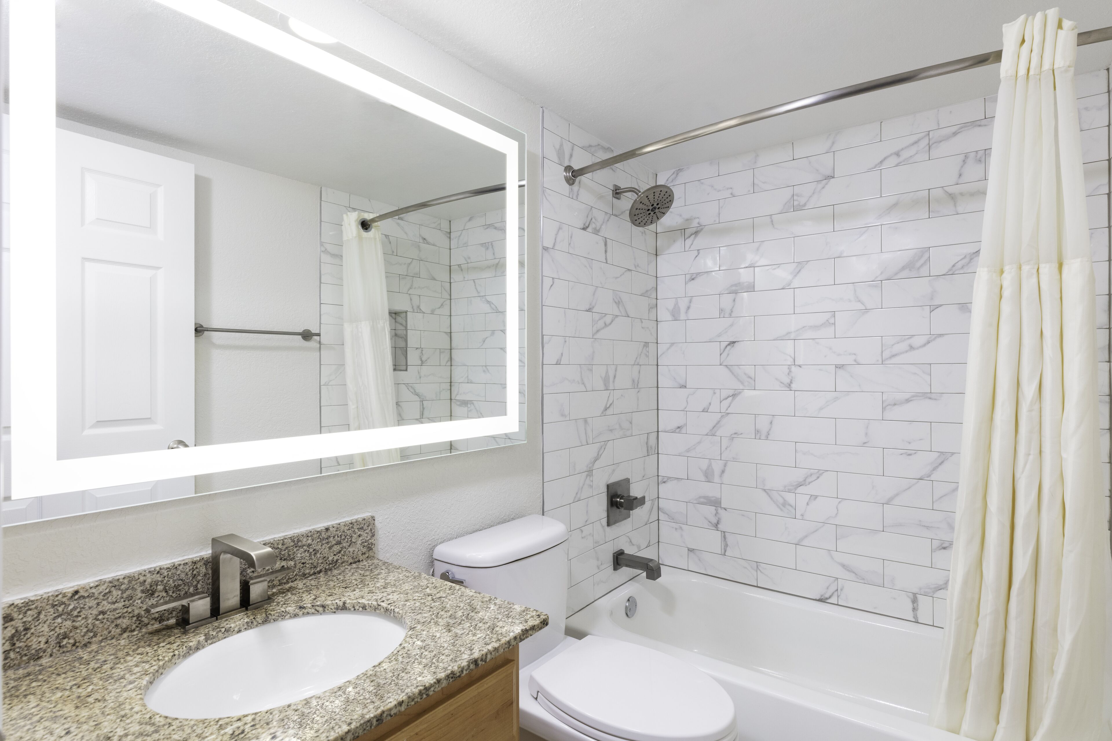 Un baño con lavamanos blanco, inodoro y bañera, paredes con diseño de mármol y un espejo grande.