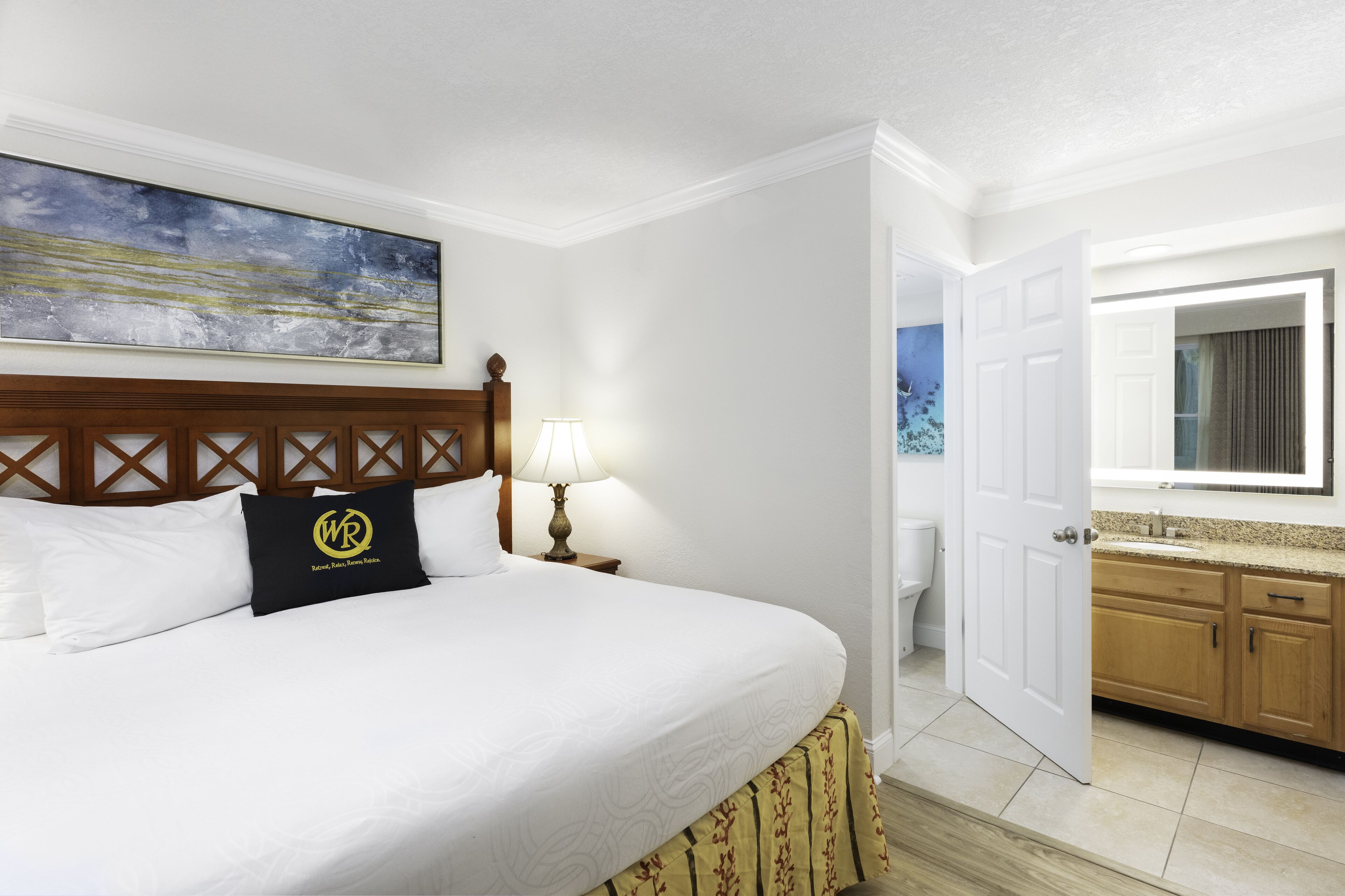 A hotel room with a large bed, a wooden headboard, a painting above the bed, a nightstand with a lamp, and a view of the bathroom through an open door.