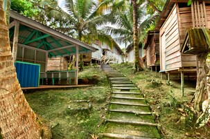 Uma escadaria de madeira estreita que sobe uma colina, cercada por palmeiras e estruturas de madeira.
