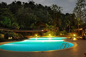 Piscina com espreguiçadeiras e área coberta, cercada por árvores densas e um edifício ao fundo.