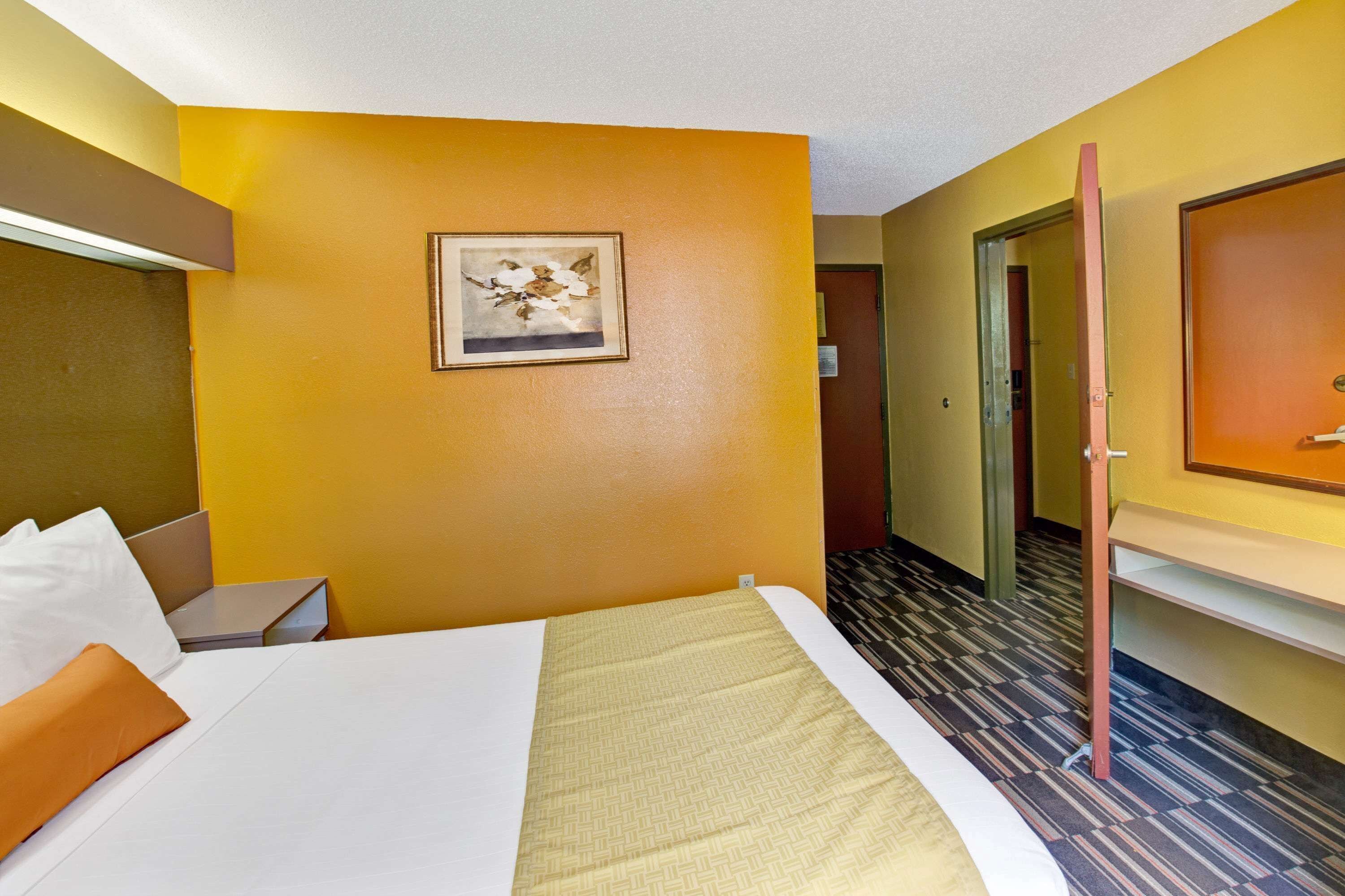 Une chambre d’hôtel avec un lit, un bureau, une chaise, un tableau encadré au mur et une porte donnant sur une autre pièce.