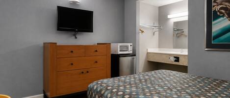Habitación de hotel con cama, cómoda, microondas, televisor y baño con espejo y toallero.