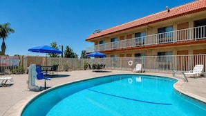 A swimming pool with lounge chairs and umbrellas, a fenced area, and a building with a red roof. - Vagabond Inn Bakersfield South (Bakersfield)