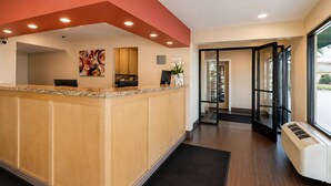 A reception area with a wooden counter, a reception desk, a wall-mounted security camera, and a door leading to another room. - Vagabond Inn Bakersfield South (Bakersfield)