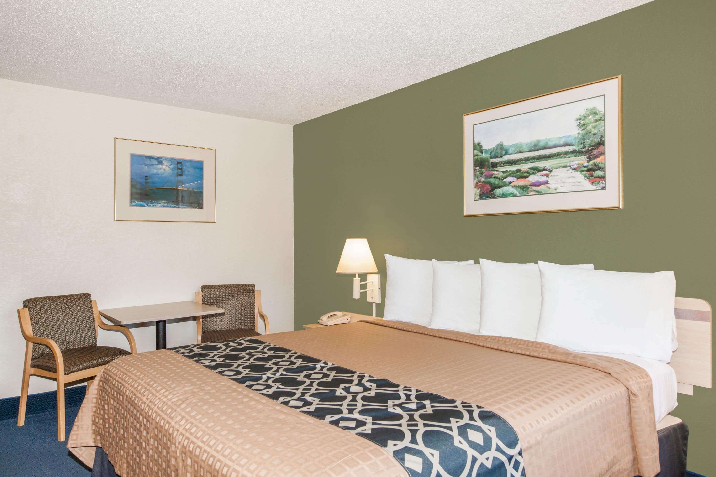 A hotel room with a bed, two chairs, a table, a lamp, and two framed pictures on the walls.