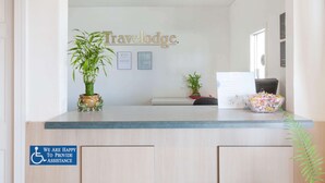 A reception desk with a sign reading "Travelodge." There are two potted plants on the desk, a framed certificate, and a sign indicating assistance for disabled guests.