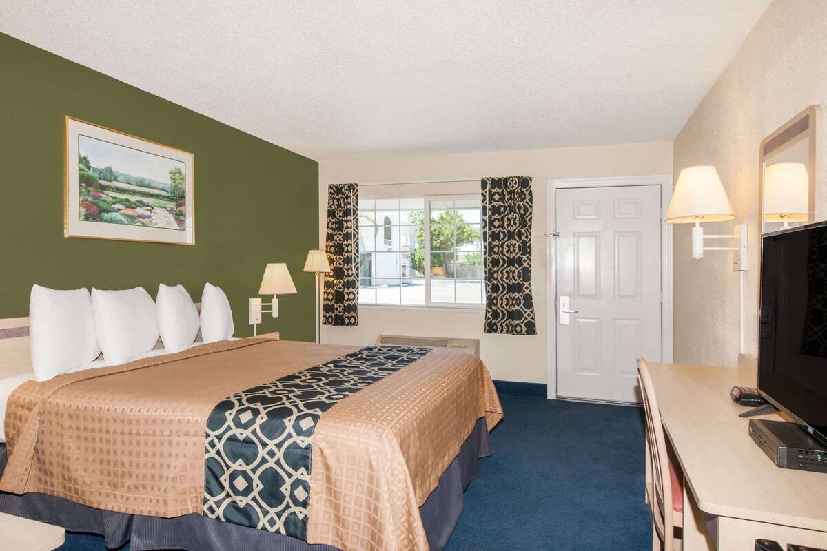 A hotel room with a large bed, a desk with a television, a window with curtains, and a painting on the wall.