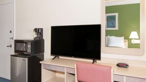 A hotel room with a desk, a television, a microwave, a refrigerator, a chair, a bed with a green wall, a bedside table with a lamp, and a digital clock.