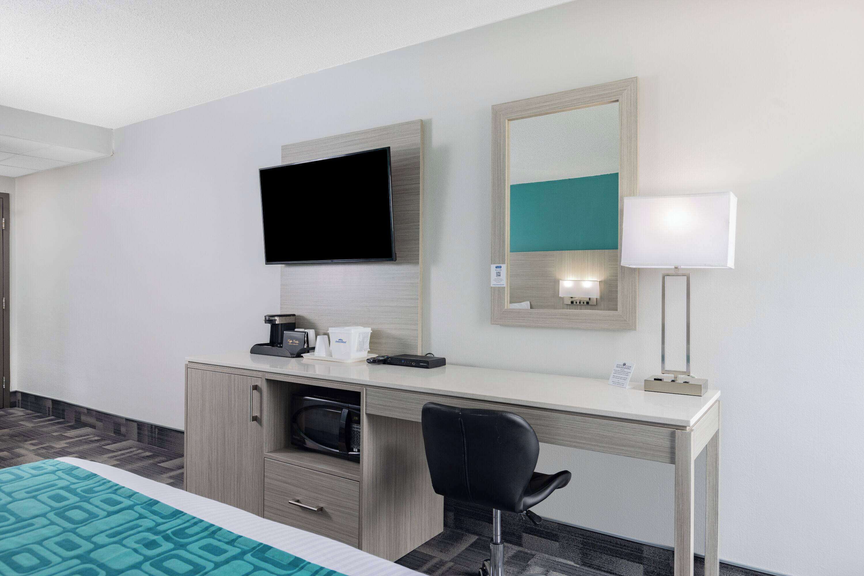 A hotel room with a bed, a desk with a flat-screen TV, a microwave, a coffee maker, a lamp, and a mirror.