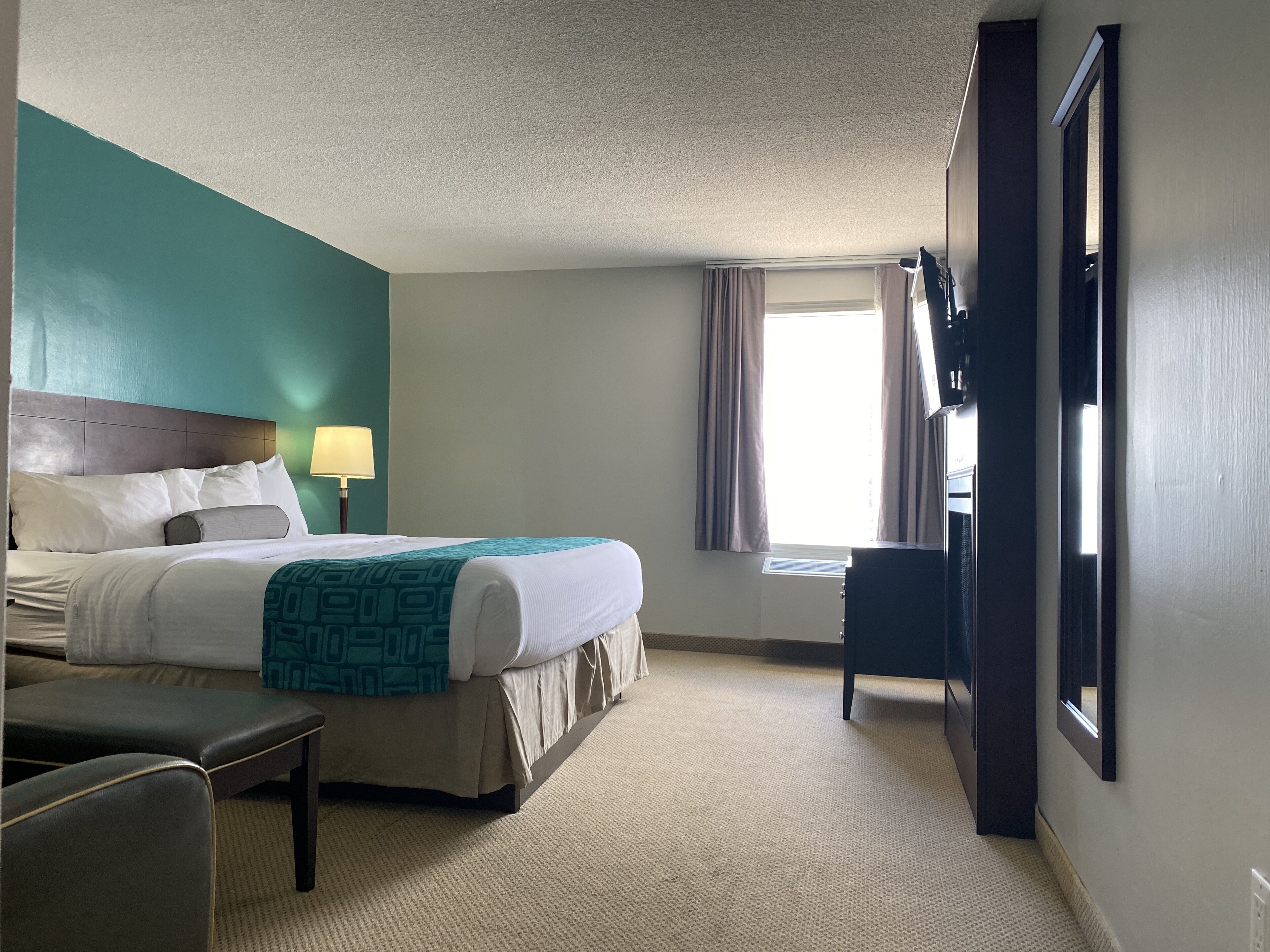 A hotel room with a bed, a desk, a chair, a lamp, a window with curtains, and a door.