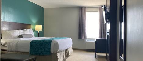 A hotel room with a bed, a desk, a chair, a lamp, a window with curtains, and a door.