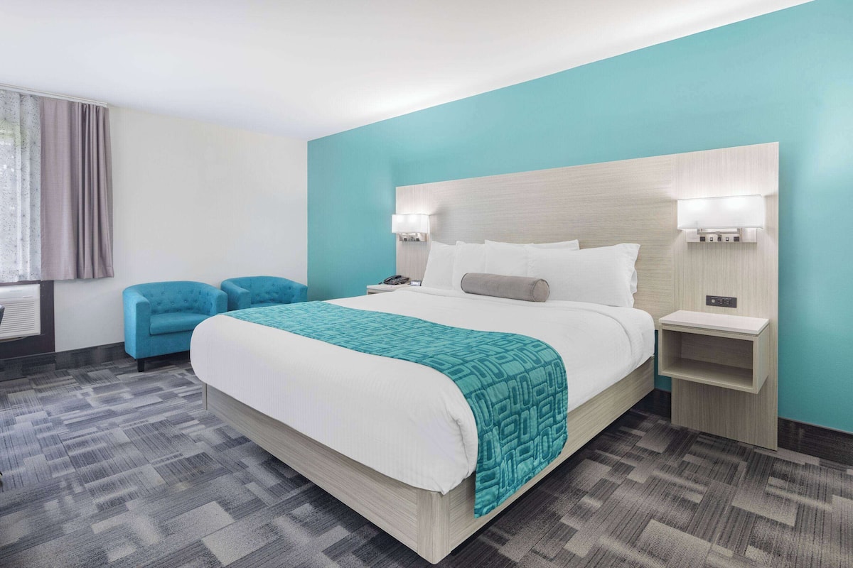 A hotel room with a large bed, two bedside tables, a nightstand, a blue armchair, and a window with curtains.