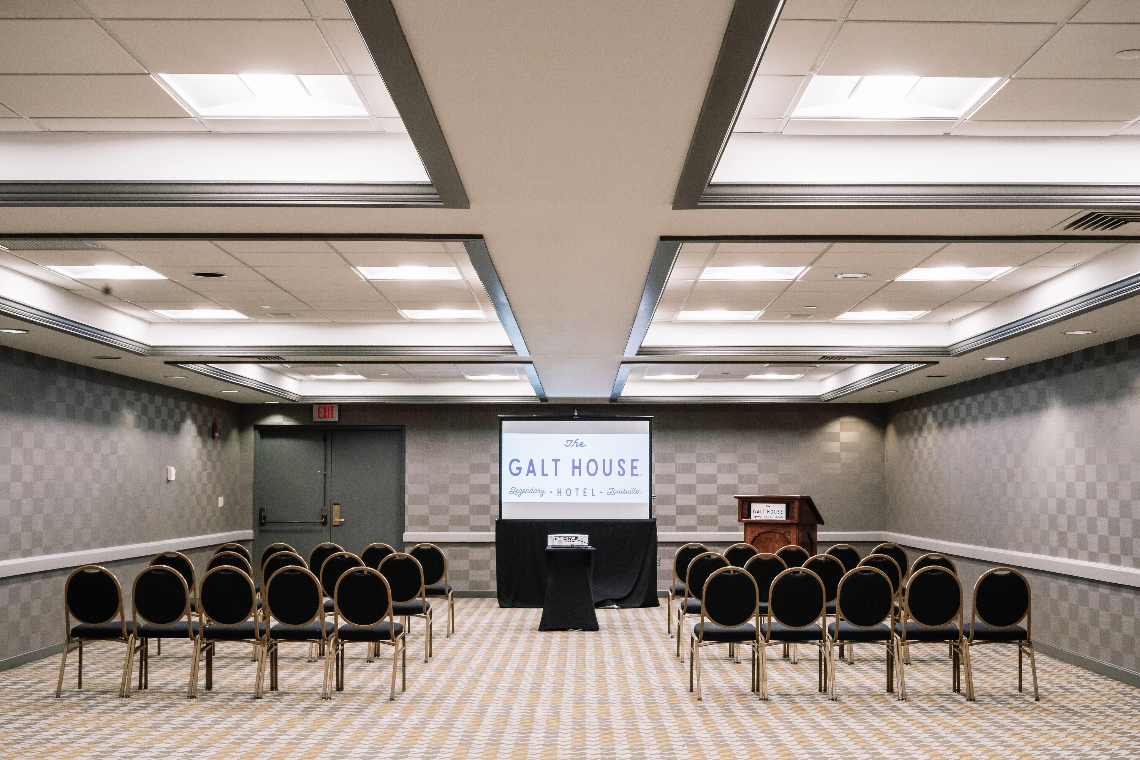 A conference room with a projector screen displaying "The Galt House Legendary Hotel Louisville" and a podium. Rows of chairs are arranged facing the screen.