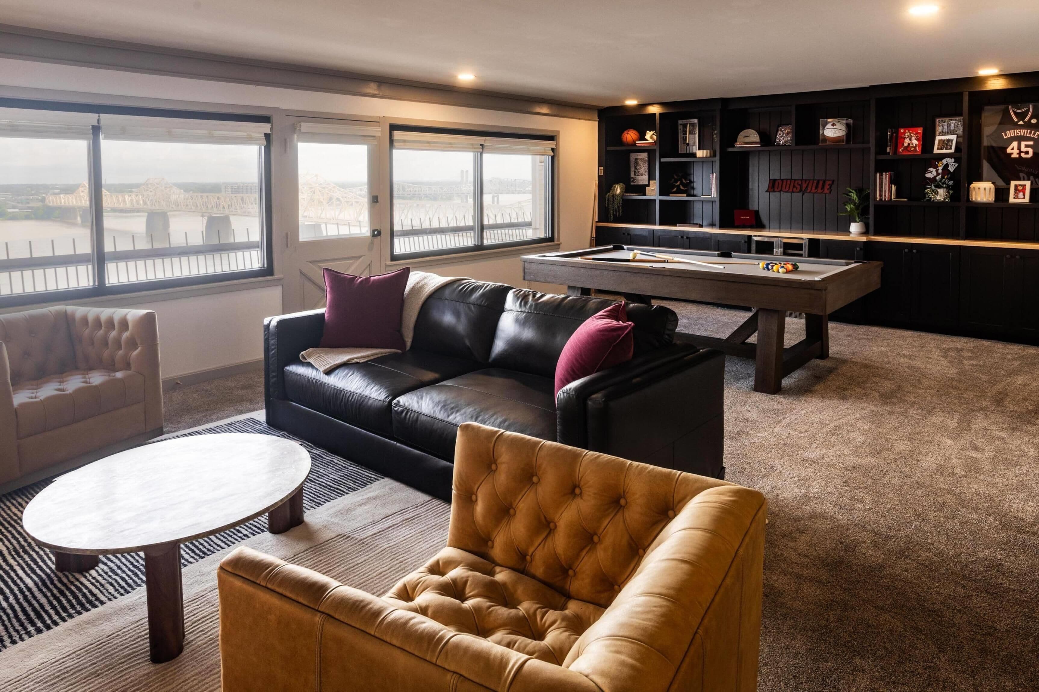 A living room with a pool table, a leather sofa, a tufted armchair, and a view through large windows.