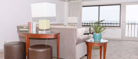 A hotel room with a sofa, two ottomans, a small table with a lamp, a potted plant, and a view of the outside through a large window.