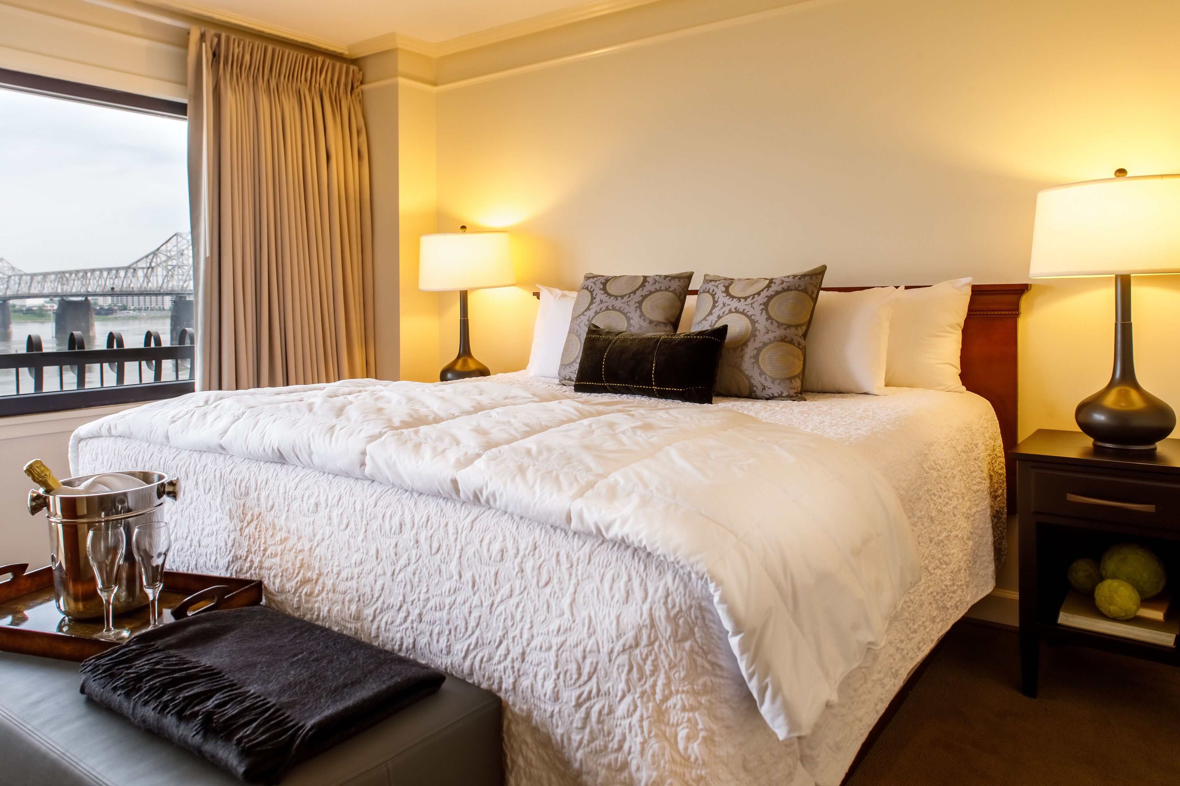 A hotel room with a large bed, two bedside lamps, a view of a bridge, and a minibar with champagne and glasses.
